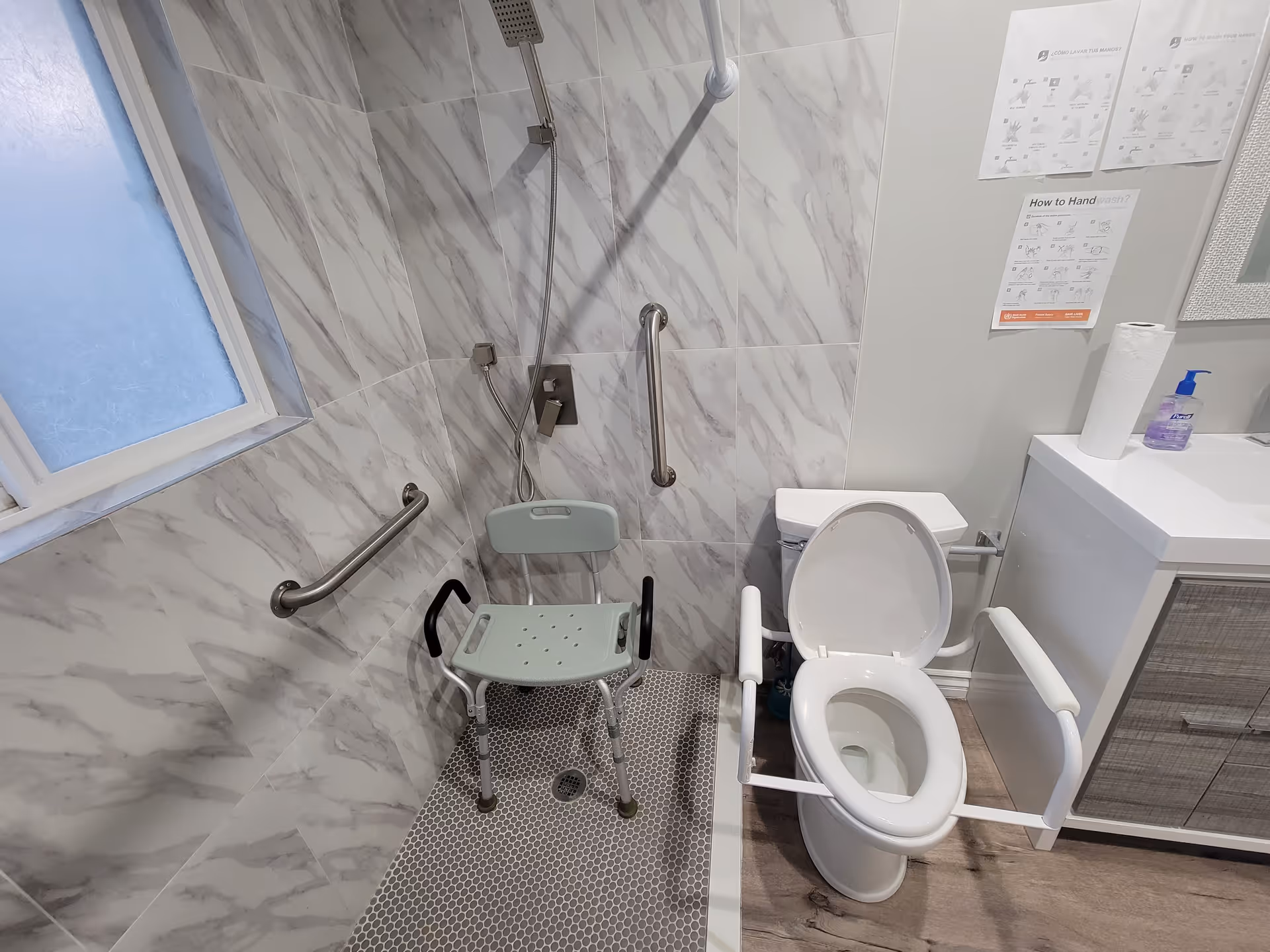 A bathroom designed for accessibility with a shower area featuring a shower chair and grab bars, a toilet with arm supports, and a sink with hand soap and paper towels. Instructions for handwashing are posted on the wall.