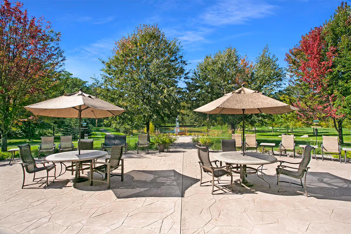 Sunlit outdoor patio with round tables, umbrellas and chairs overlooking a green lawn, trees, and a distant fountain.