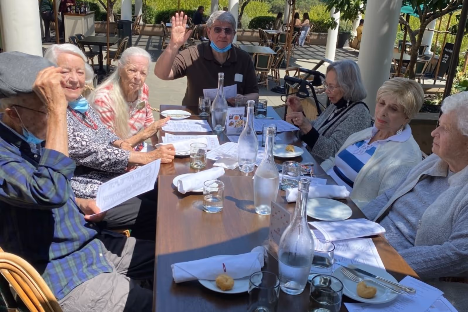A group of elderly people sitting around a long outdoor table at a senior living facility, looking at menus and engaging with each other. One man is raising his hand, and there are water bottles, glasses, and plates on the table. The setting is a shaded patio area with greenery in the background.
