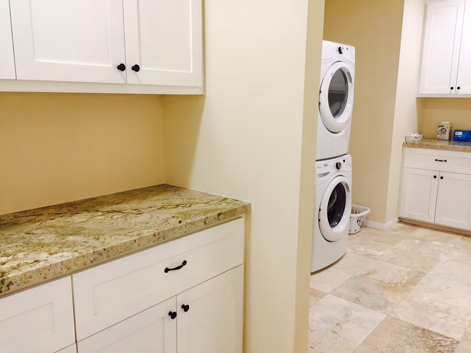 Laundry room with white cabinets, granite countertops, and a stacked washer and dryer. The floor is tiled with beige stone tiles.