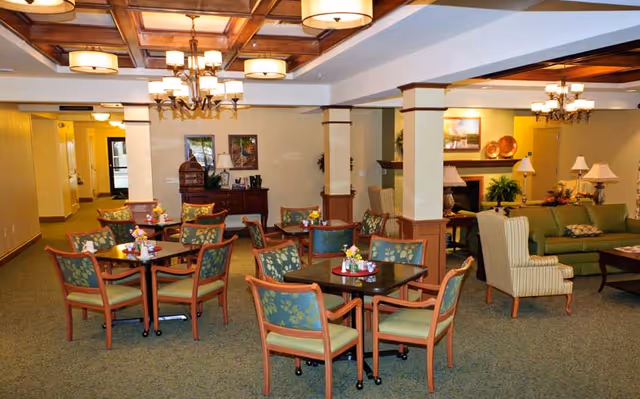 A spacious common area with multiple small square tables surrounded by wooden chairs with green floral cushions. The room features a coffered ceiling with several light fixtures, beige walls with wooden trim, and a carpeted floor. In the background, there is a seating area with green sofas, armchairs, lamps, and a fireplace with decorative plates on the mantel.