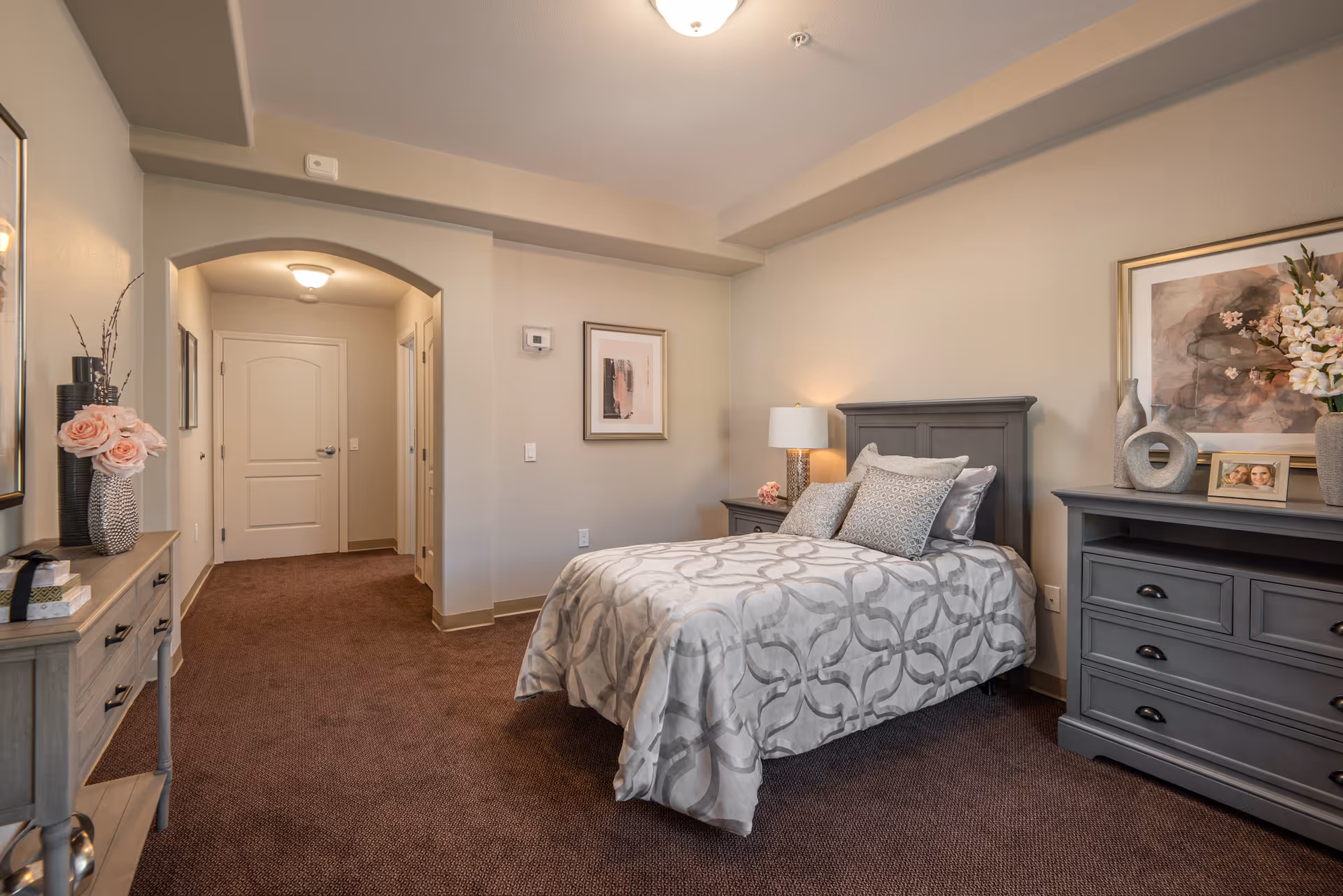 A neatly arranged bedroom in a senior living facility with a single bed covered in a patterned gray and white comforter, two gray pillows, a nightstand with a lamp and flowers, a dresser with decorative vases and a framed photo, beige walls, brown carpet, and an arched doorway leading to a hallway with closed doors.