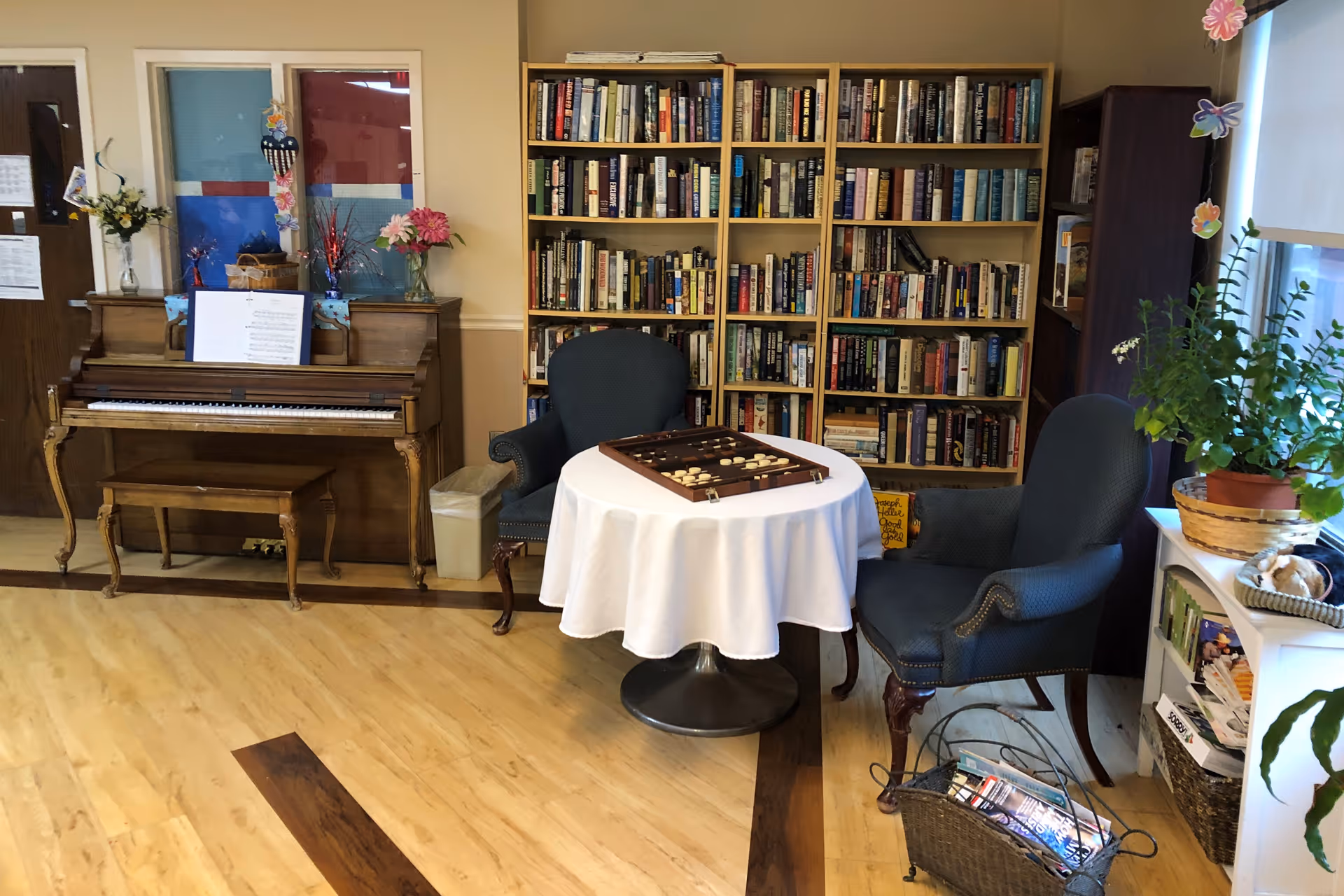 A cozy room with a wooden piano on the left, a small round table covered with a white tablecloth in the center, two upholstered armchairs on either side of the table, and a bookshelf filled with books against the back wall. There are flowers on the piano and a potted plant on a white shelf near a window with a roller shade. A backgammon game is set up on the table.