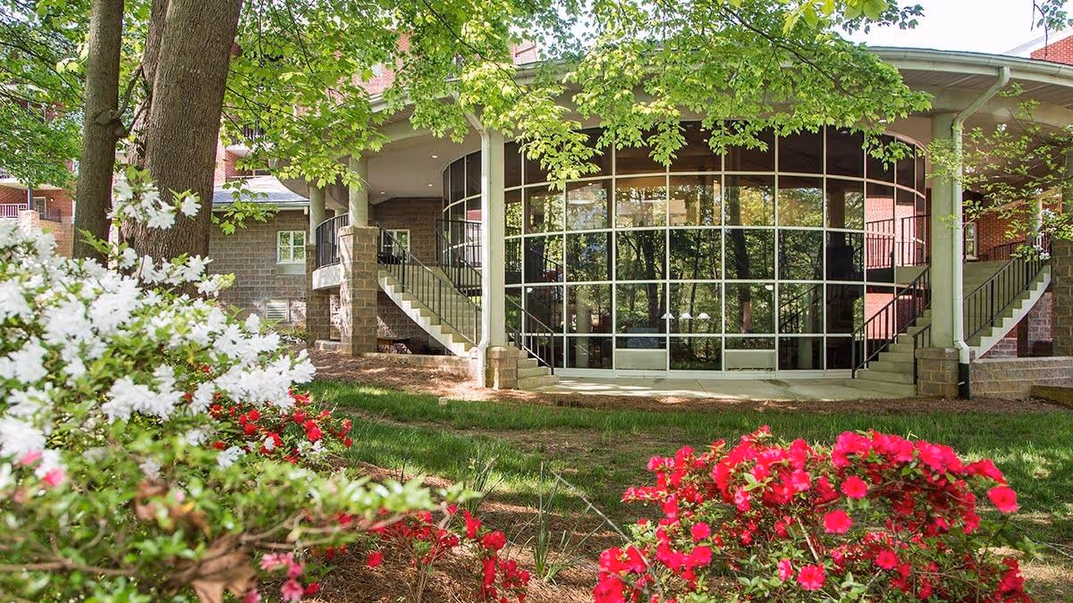 Exterior view of Brookridge Life Plan Community showing a building with large curved glass windows, two staircases on either side, surrounded by green trees and colorful flowering bushes in the foreground.