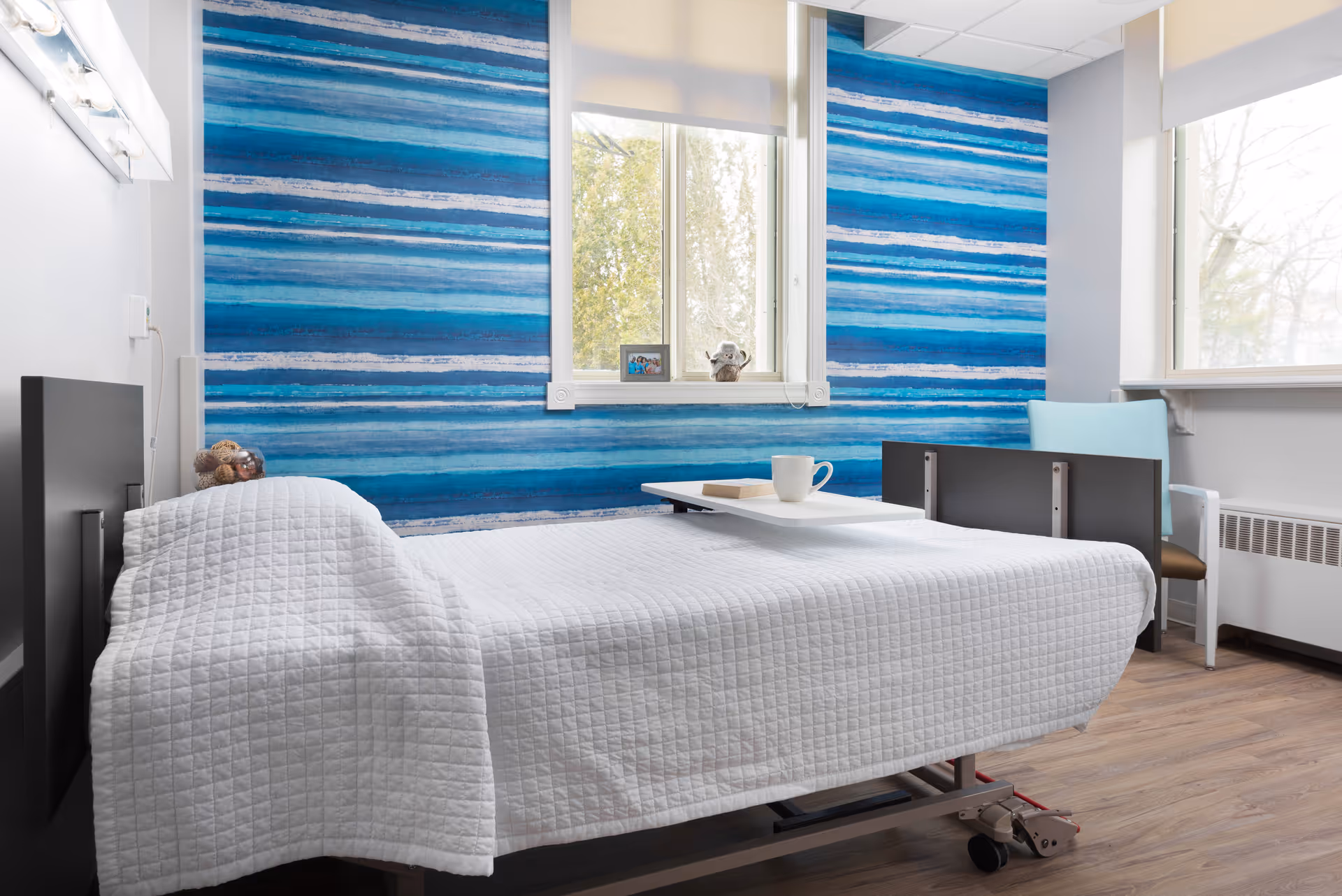 A bright and clean bedroom in a healthcare facility featuring a hospital bed with a white quilted blanket, a small overbed table with a cup and a book, a blue and white striped accent wall behind the bed, a window letting in natural light, and a light blue chair next to the bed.