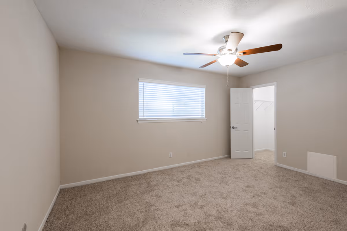 Empty bedroom with beige carpet, beige walls, a window with blinds, a ceiling fan with light, and an open door leading to a walk-in closet.