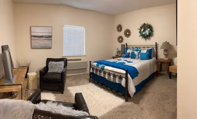 A cozy bedroom with a metal frame bed dressed in white and teal bedding, decorative pillows, and a floral wreath above the headboard. The room features beige walls, a window with blinds, a black armchair with a gray pillow, a wooden TV stand with a television, a side table with a lamp, and carpeted flooring.