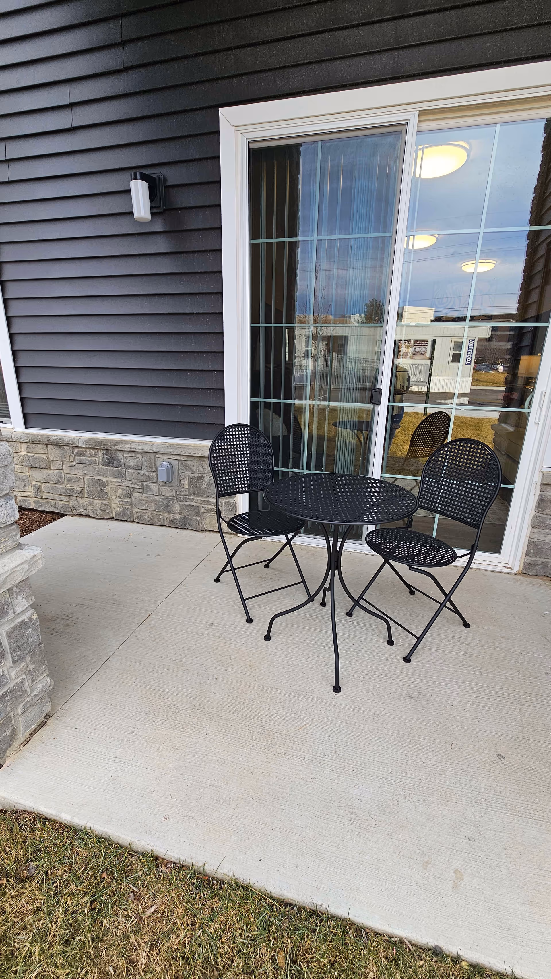 Outdoor patio area with a small round black metal table and two matching black metal chairs on a concrete floor. The patio is adjacent to a building with dark siding and stone accents, and there is a sliding glass door with vertical blinds behind the table and chairs.