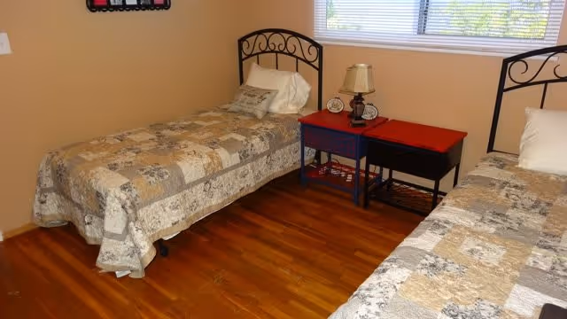 A bedroom with two twin beds featuring metal headboards and patchwork quilts in neutral tones. Between the beds is a small table with a lamp and a clock. A window with blinds lets in natural light, and the floor is wooden.