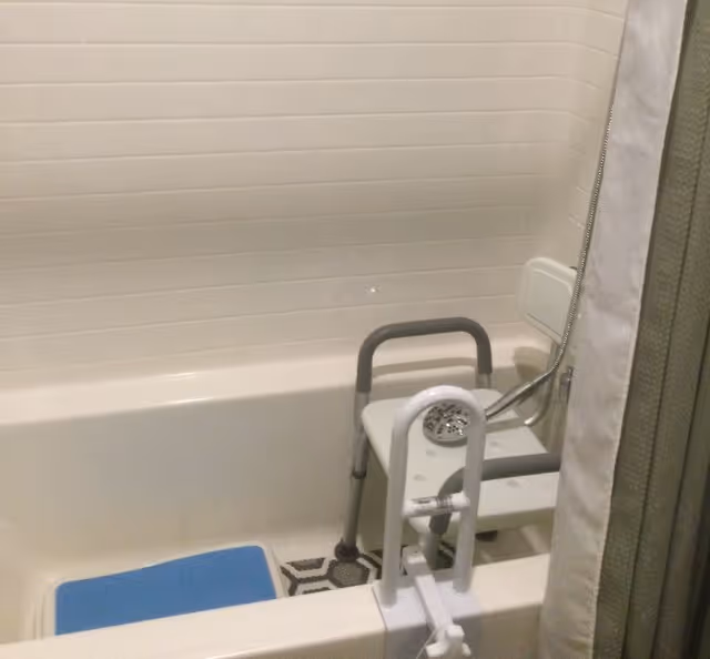 A bathtub with white tiled walls, equipped with a white shower chair with gray handles and a handheld showerhead. There is a blue bath mat inside the tub and a white safety grab bar attached to the tub edge.