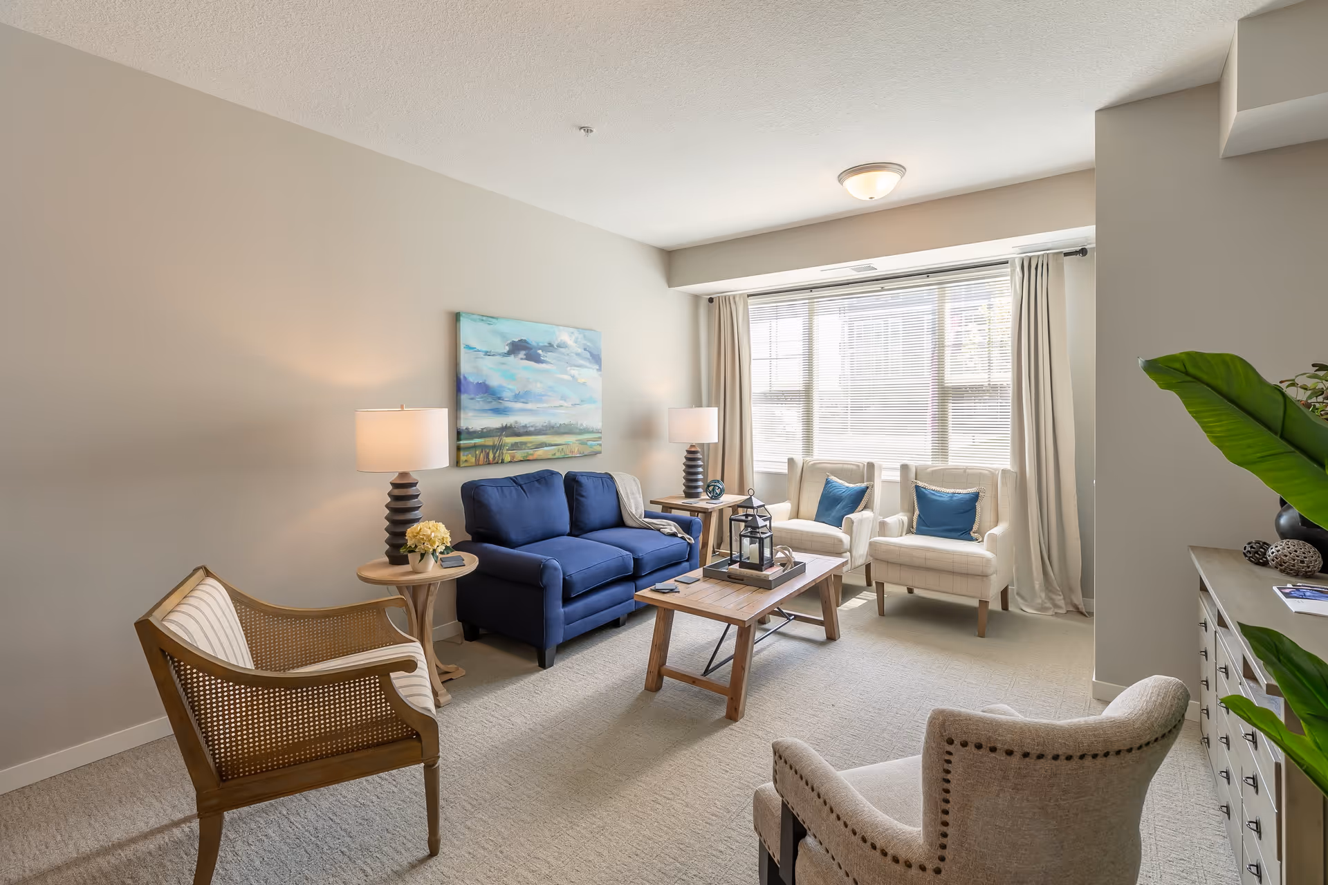 A bright and cozy living room with a blue loveseat, two beige armchairs with blue pillows, a wooden coffee table, a wooden chair with cane sides, two side tables with lamps, a large window with blinds and curtains, a painting on the wall, and a plant on a dresser.