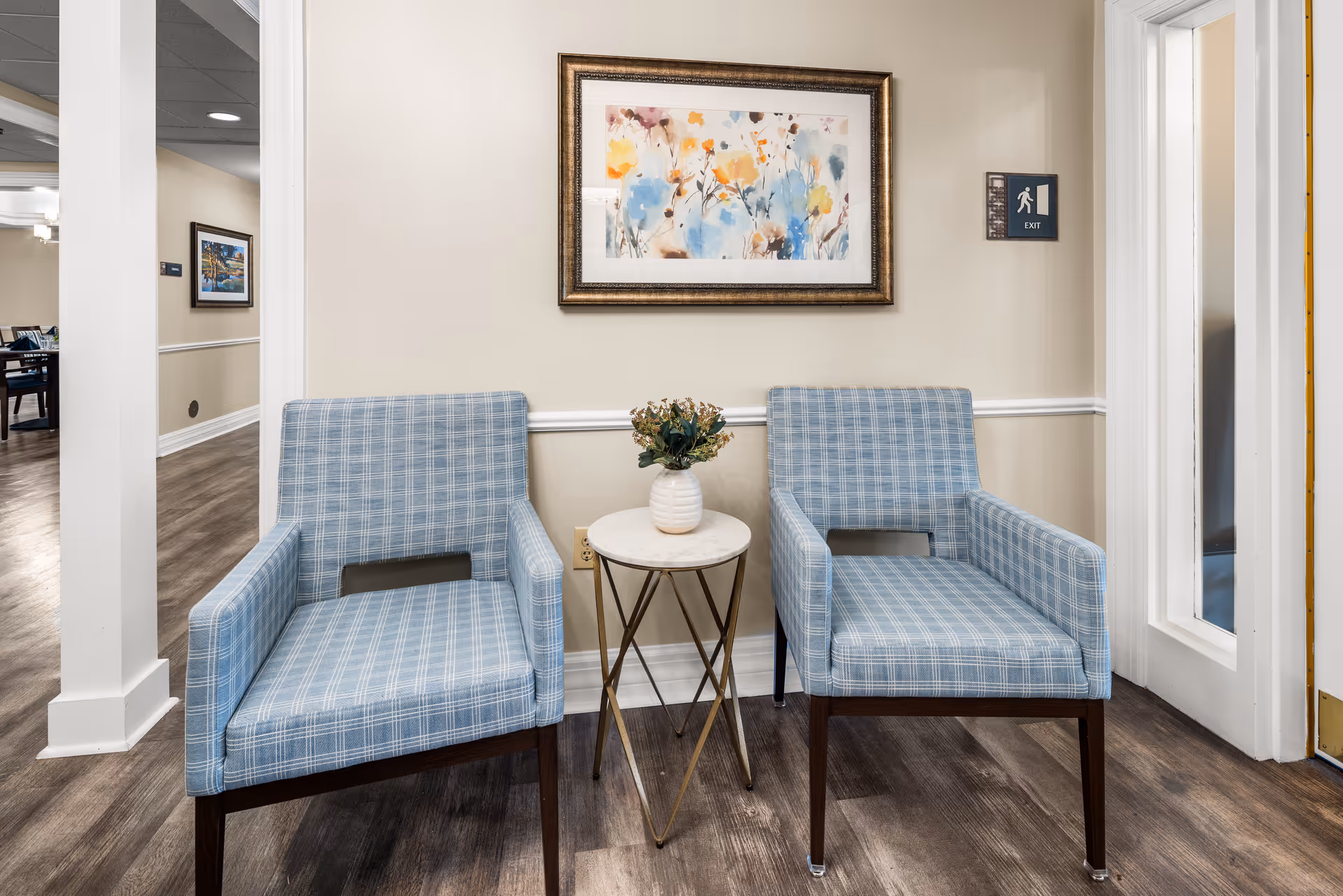 Two blue plaid armchairs with wooden legs are positioned on either side of a small round table with a white marble top and gold metal legs. A small white vase with green and brown foliage sits on the table. Behind the chairs, a beige wall features a framed abstract floral painting with colors including yellow, orange, blue, and brown. To the right, there is a door with glass panels and an exit sign above it. The floor is a dark wood laminate, and an adjacent room with dining tables and chairs is visible through an open doorway on the left.