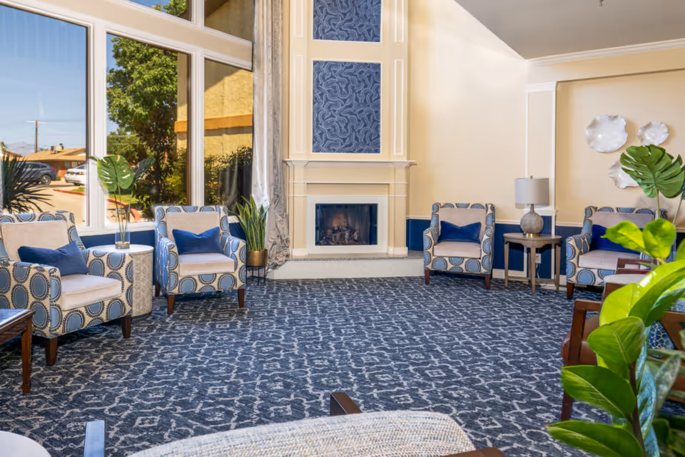 Bright seating lounge with patterned armchairs, a fireplace, large windows, and houseplants.