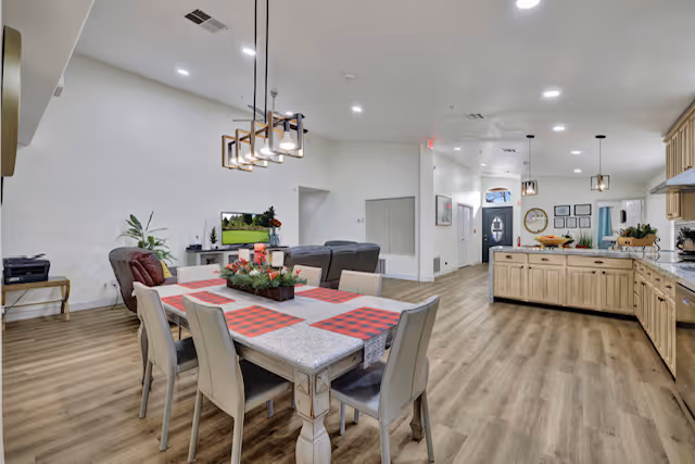 A spacious, open-concept interior of a senior living facility showing a dining area with a table set with red and black checkered placemats and a centerpiece, adjacent to a living room with a TV and seating, and a kitchen with wooden cabinets and a large island. The flooring is wood-style, and the walls are painted white with recessed lighting overhead.