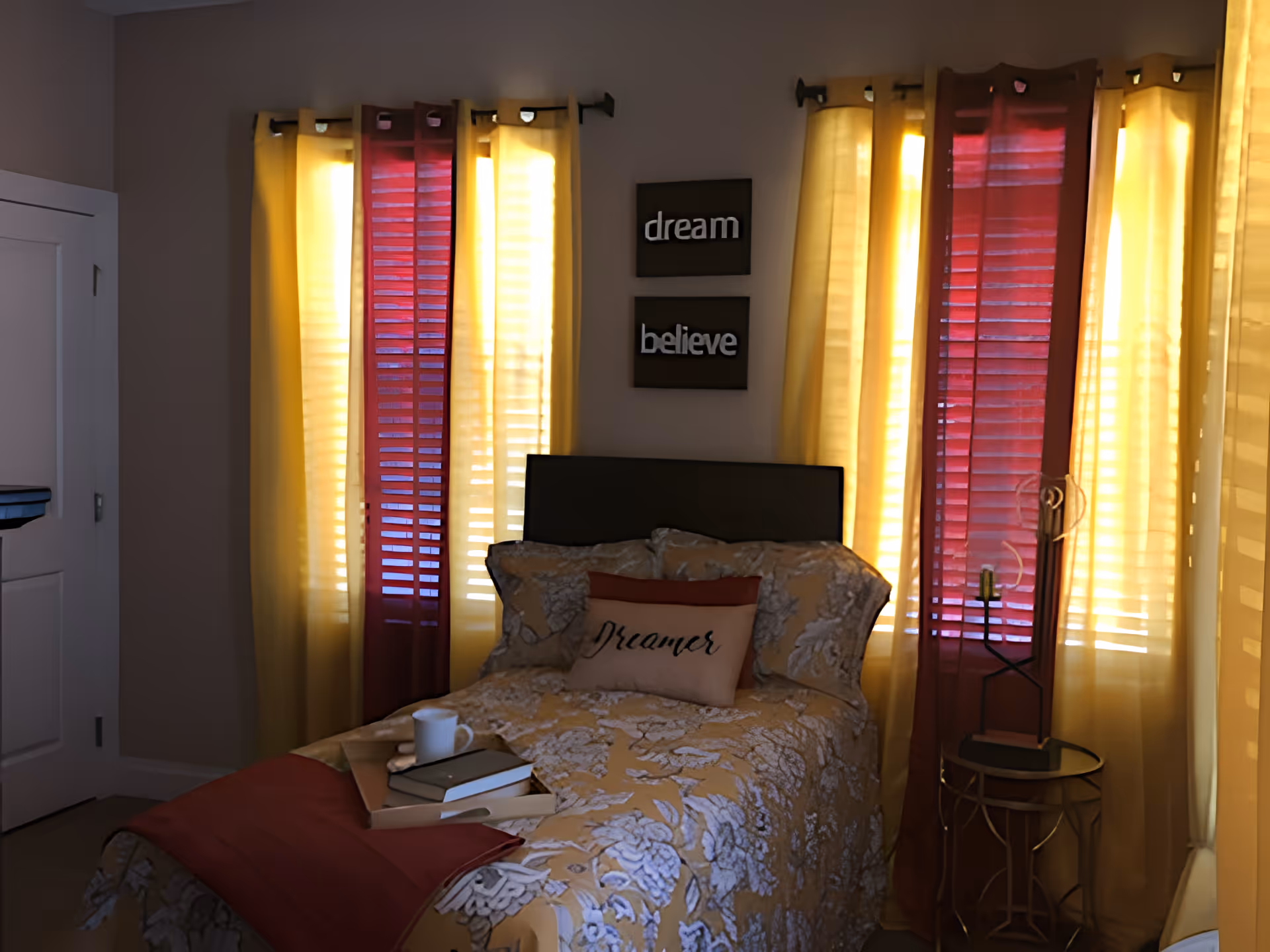 A cozy bedroom with a bed covered in a floral patterned bedspread and pillows, including one with the word 'Dreamer'. The bed is positioned in front of windows with yellow and red curtains. Above the bed, two wall plaques read 'dream' and 'believe'. A small round side table with a candle holder is next to the bed. A tray with books and a cup is placed on the bed.
