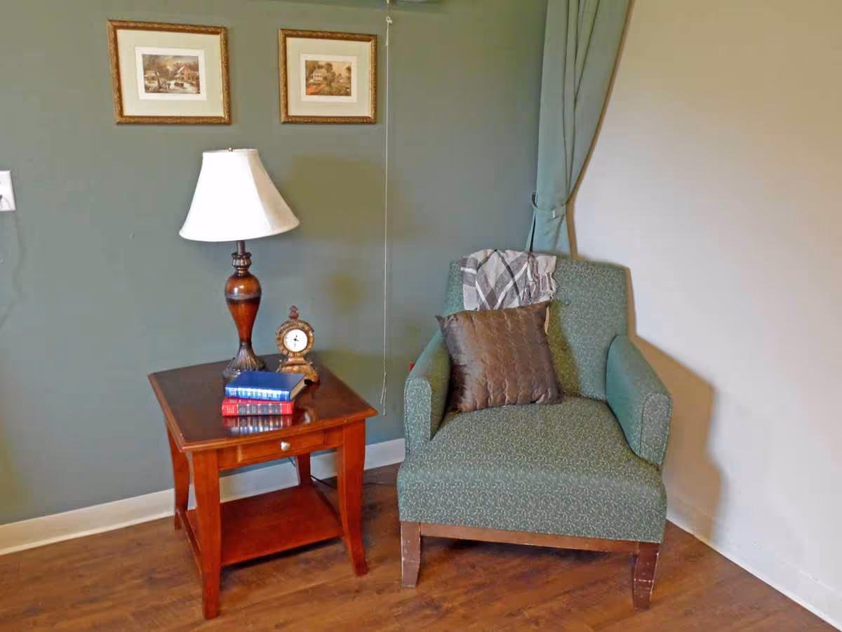 A cozy corner with a green upholstered armchair that has a brown pillow and a gray throw draped over the back. Next to the chair is a wooden side table with a drawer, holding a table lamp with a white shade, a small vintage clock, and a stack of three books. The wall behind is painted green and decorated with two framed pictures. The floor is wooden.