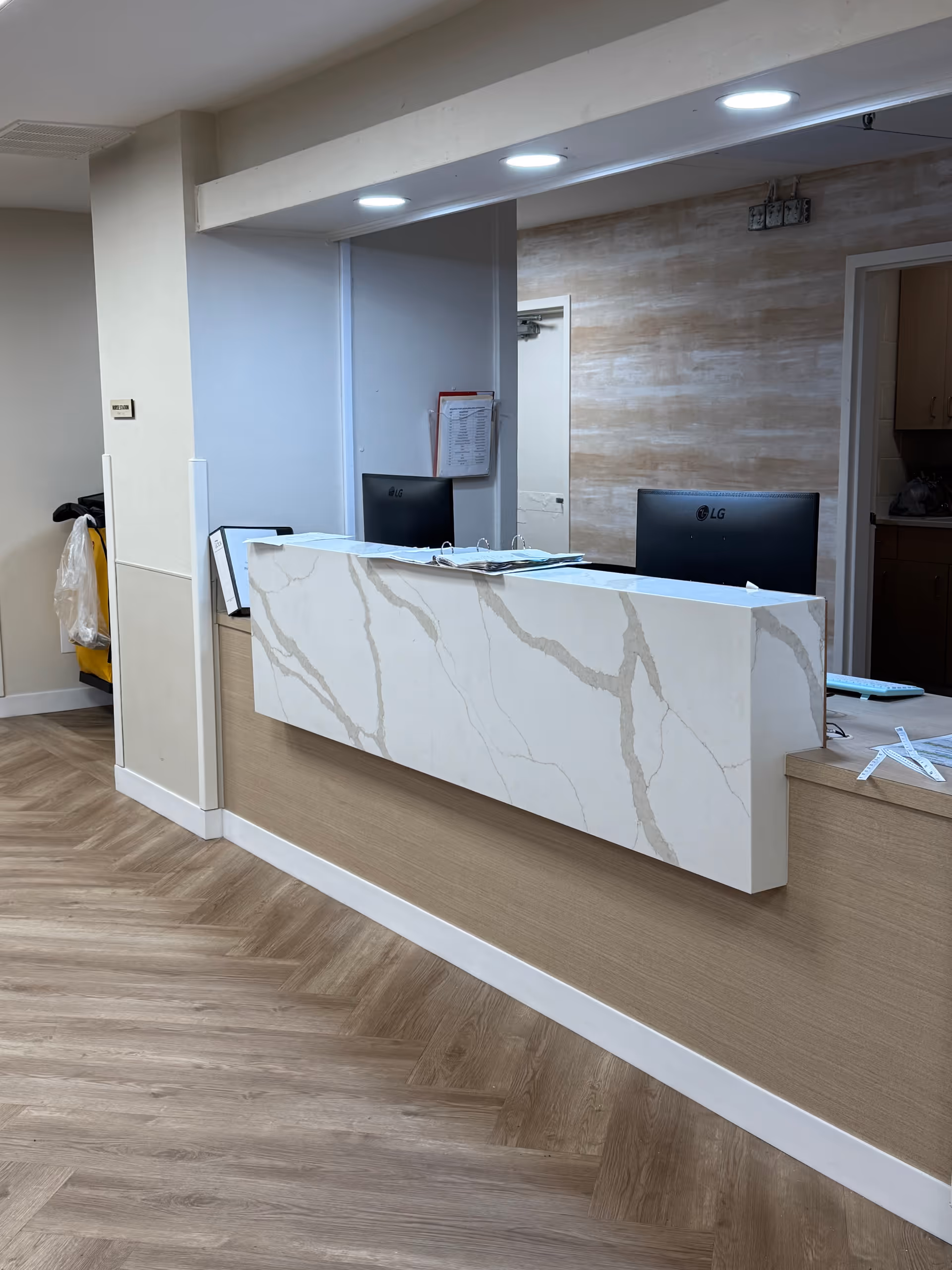 Reception desk area with a marble-patterned front panel, two computer monitors, and some papers on the counter. The floor has a light wood pattern, and the walls are painted in neutral tones. There is a doorway behind the desk and some cleaning supplies visible to the left.