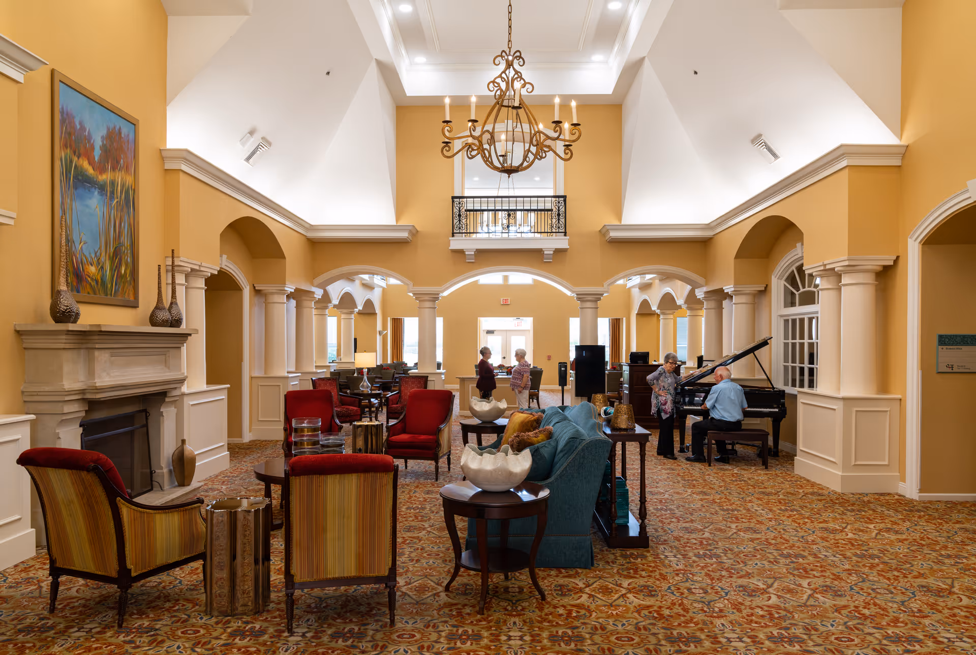 A spacious and elegant common area in a senior living facility with high vaulted ceilings, a large chandelier, and warm yellow walls. The room features a fireplace with a painting above it, several upholstered chairs and sofas arranged around small tables, and a grand piano being played by a man while a woman stands nearby. Two other people are seen conversing in the background near the entrance.