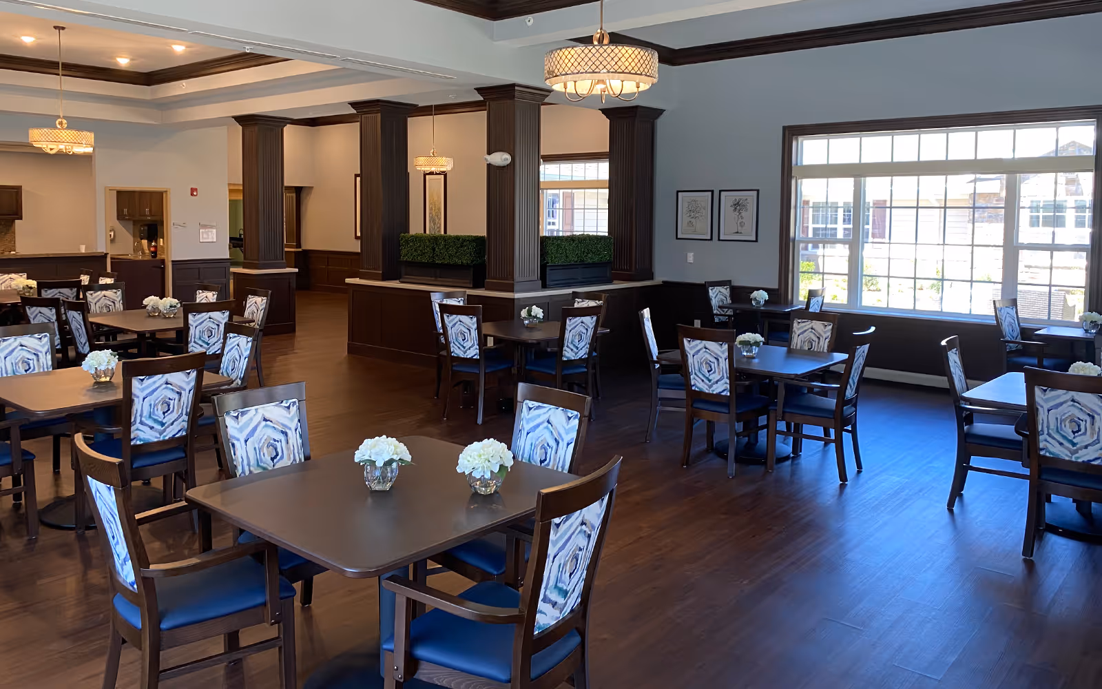 A spacious dining room with multiple dark wooden tables and chairs featuring patterned upholstery. Each table has a small floral centerpiece. Large windows on one side let in natural light, and decorative pendant lights hang from the ceiling. The room has wooden flooring and dark wood paneling on the lower walls.