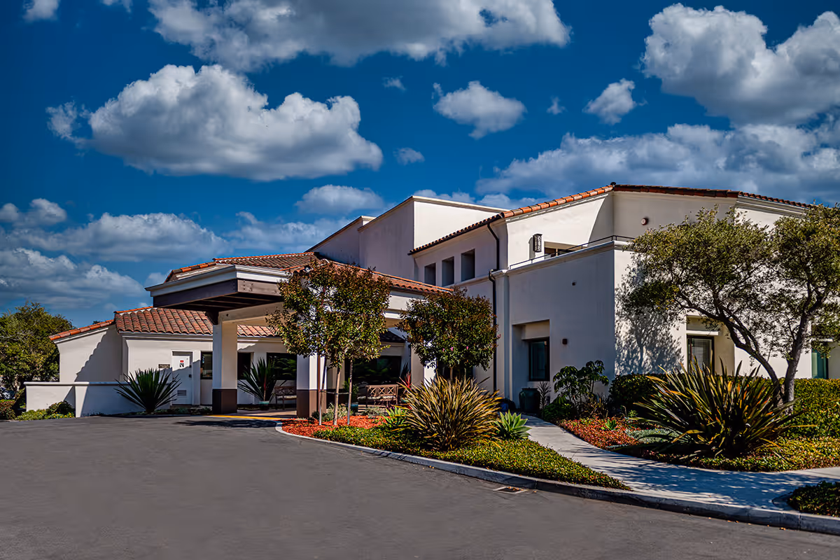 Mediterranean-style senior living building front with a covered entrance, driveway, and landscaped grounds under a blue sky.