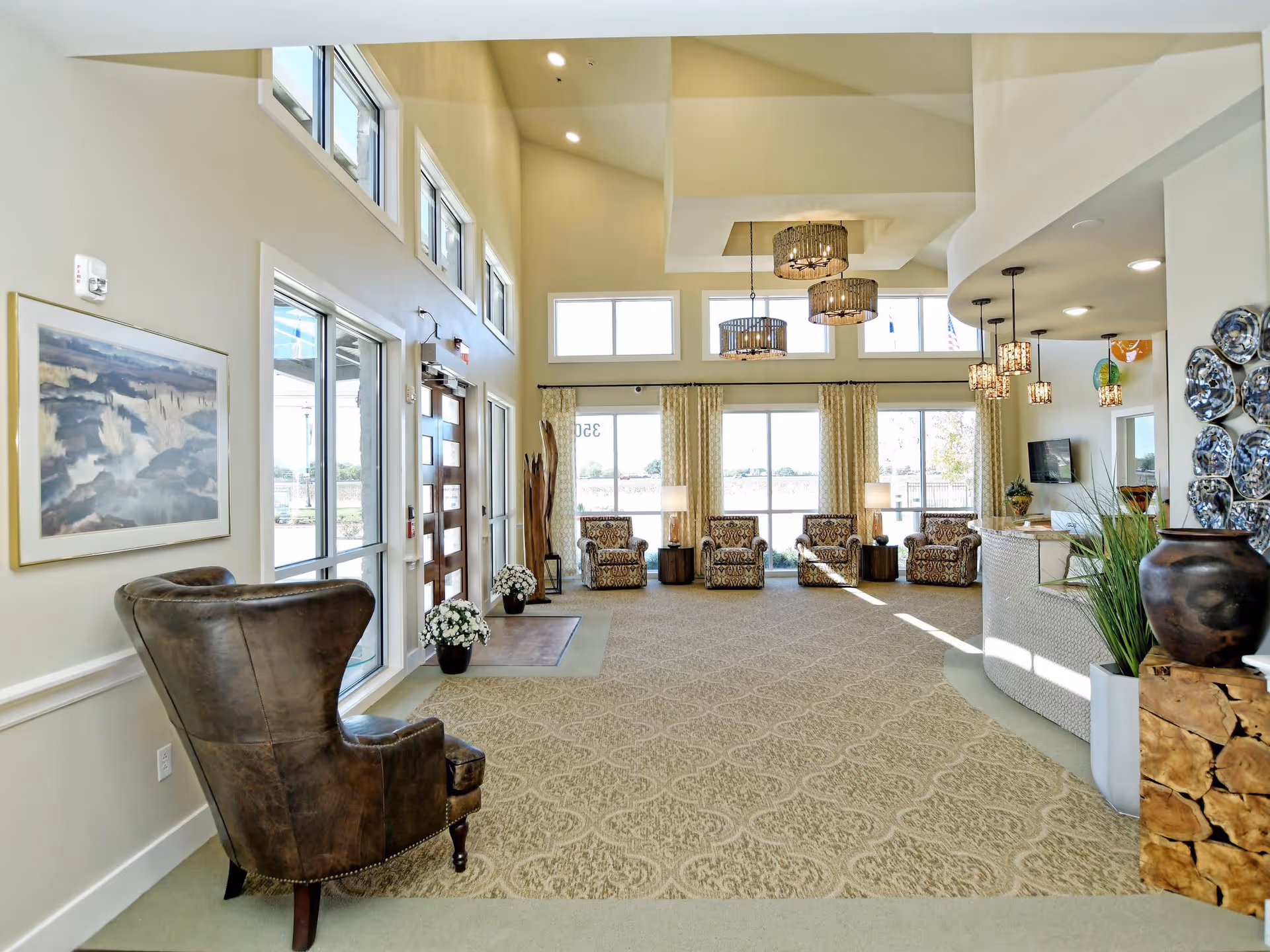 A spacious, well-lit common area with high ceilings and large windows. The room features a patterned carpet, a dark leather armchair in the foreground, and a row of four patterned armchairs along the far wall with side tables and lamps. There are modern pendant lights hanging from the ceiling, a reception desk on the right with decorative items including a large vase and a plant, and a wall adorned with decorative plates. The entrance door has glass panels and there are framed artworks on the walls.