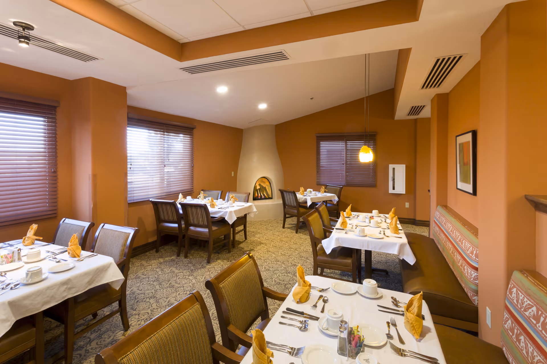 A cozy dining room with several tables set with white tablecloths, folded yellow napkins, cups, plates, and silverware. The room has warm orange walls, windows with blinds, a corner fireplace, and cushioned bench seating along one wall.
