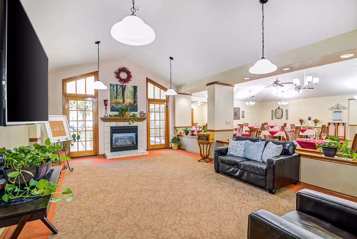 A bright and spacious common area in a senior living facility featuring a cozy seating area with black leather couches and blue cushions, a fireplace with a painting and wreath above it, large windows and glass doors letting in natural light, and a dining area with tables covered in red tablecloths and wooden chairs in the background. Several hanging pendant lights and ceiling fans illuminate the space, and there are various potted plants adding greenery.