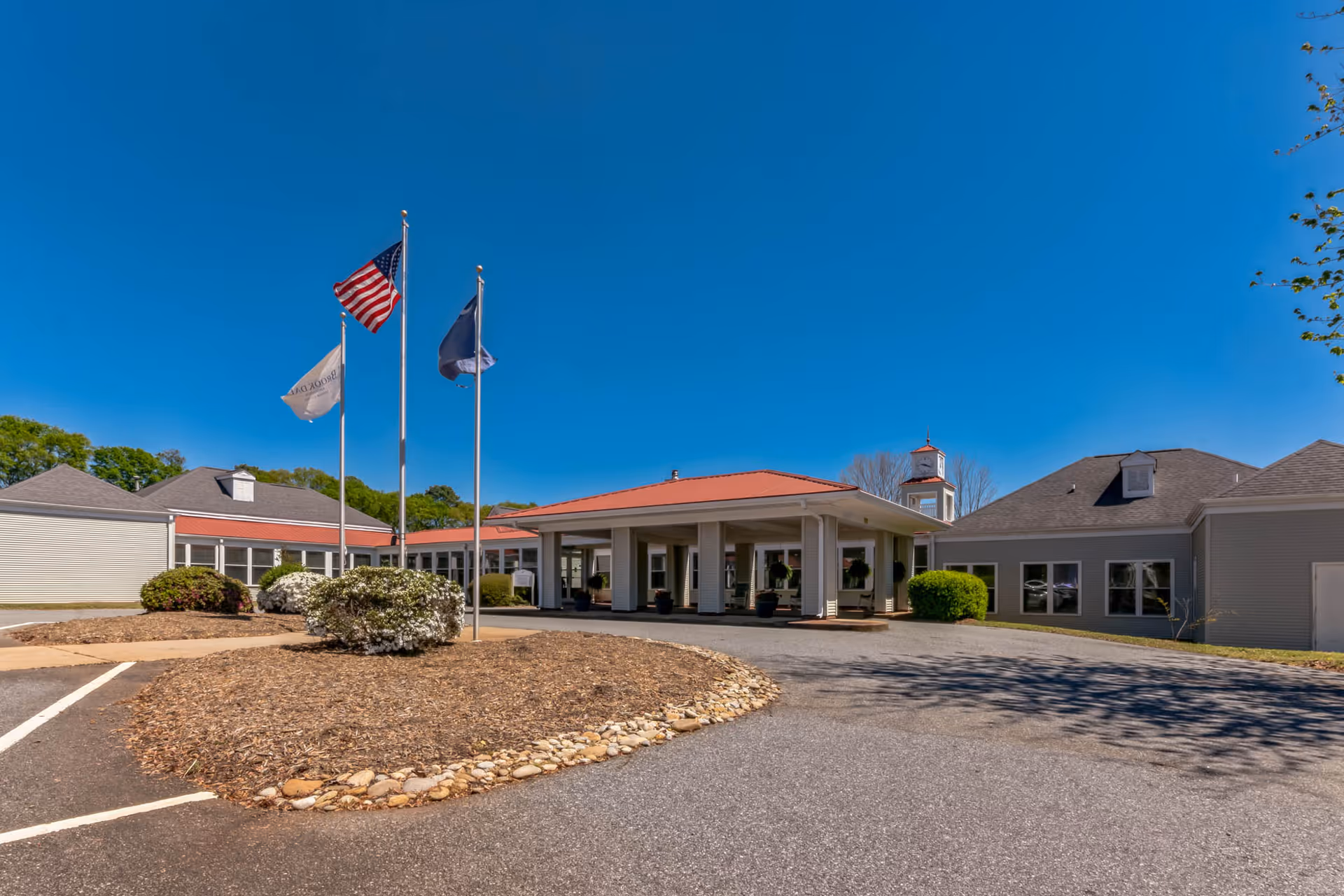 Front entrance of a single-story senior living building with a covered porte-cochère, three flagpoles, and a circular landscaped driveway under a clear blue sky.