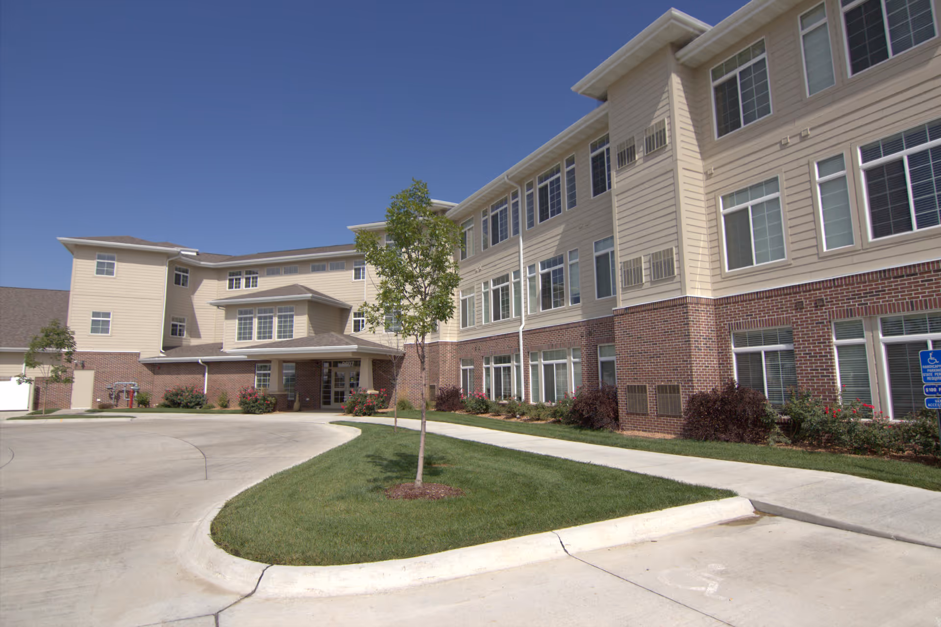 Exterior view of a three-story senior living facility building with beige siding and red brick accents. The building has many large windows and a covered entrance. There is a curved driveway and a small landscaped area with a young tree and bushes. The sky is clear and blue.