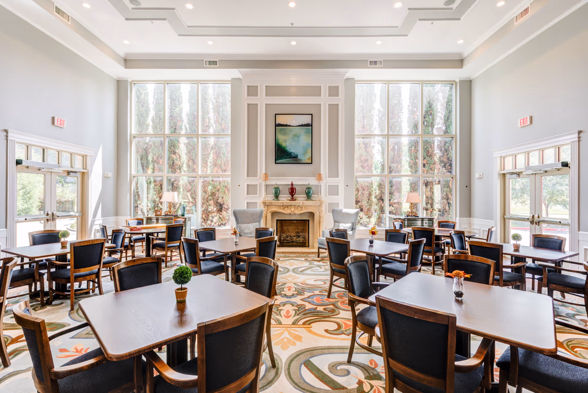 A bright and spacious dining room with multiple wooden tables and chairs arranged neatly. The room features large floor-to-ceiling windows allowing natural light to flood in, with tall trees visible outside. There is a decorative fireplace in the center of the far wall, flanked by two armchairs and small tables with lamps. The carpet has a colorful, intricate pattern, and small potted plants and flowers adorn the tables.