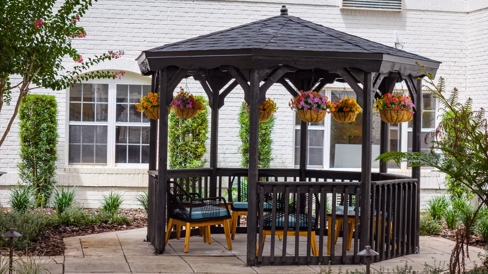 A black wooden gazebo with a shingled roof in a garden courtyard. The gazebo has hanging baskets filled with colorful flowers and contains several chairs with blue cushions. Surrounding the gazebo are green plants and shrubs, with a white brick building and windows in the background.