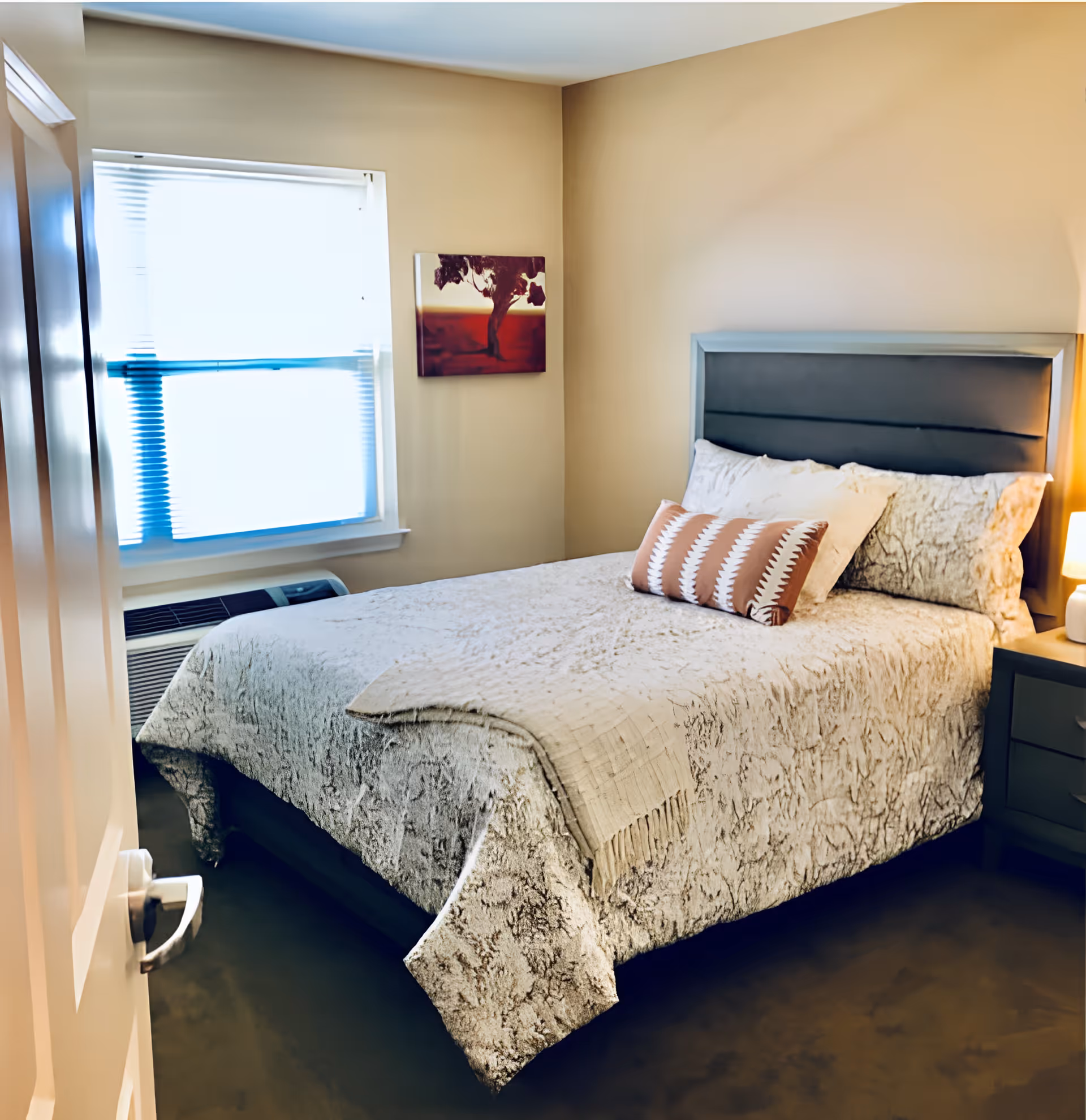A cozy bedroom with a neatly made bed featuring a textured bedspread and decorative pillows. There is a window with blinds partially open, a small nightstand with a lamp, and a piece of artwork depicting a tree on the wall.