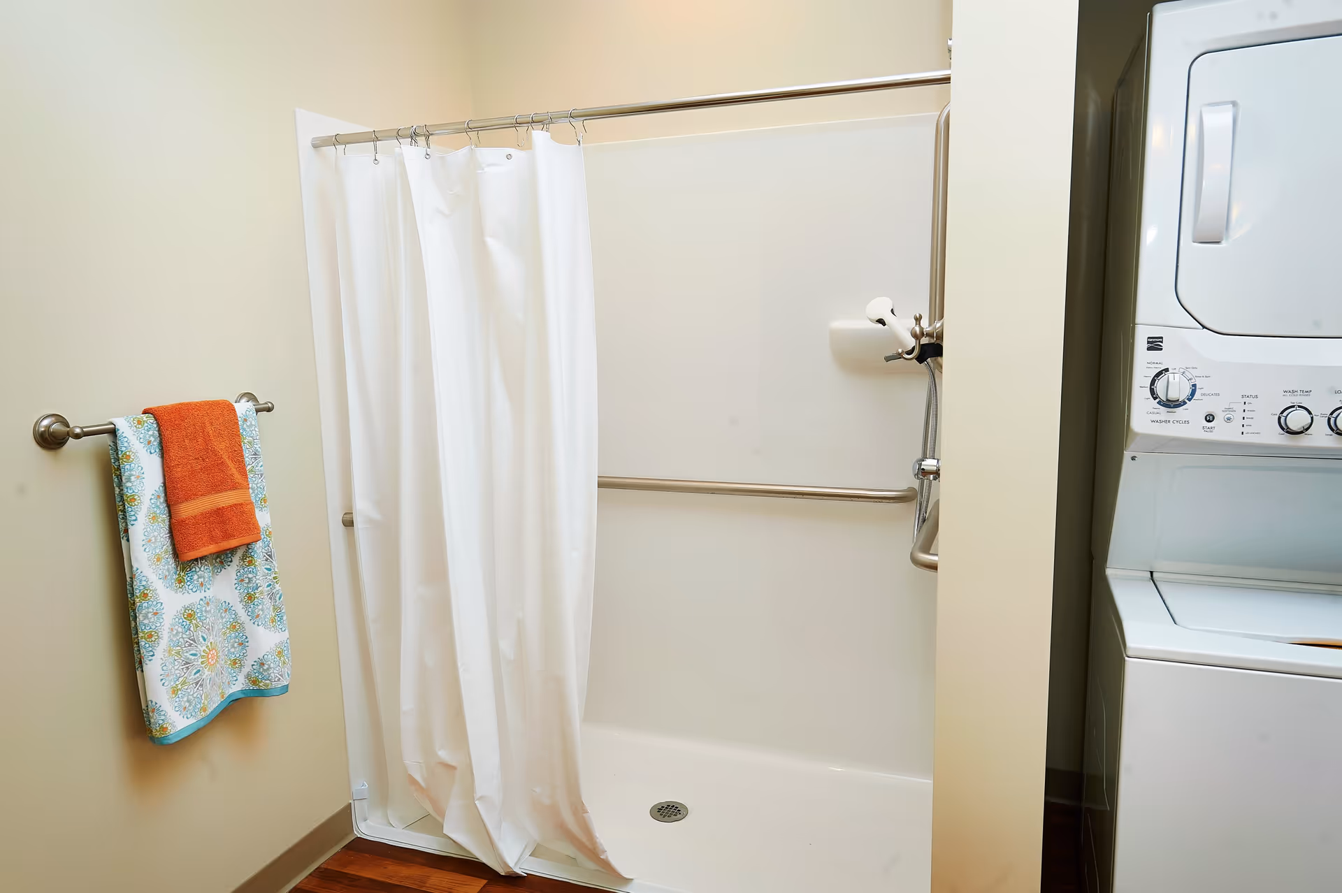 A bathroom with a white shower area featuring a white shower curtain and a metal grab bar. To the left, there is a towel rack with two towels, one orange and one patterned with blue and green designs. On the right side, there is a stacked white washer and dryer unit.