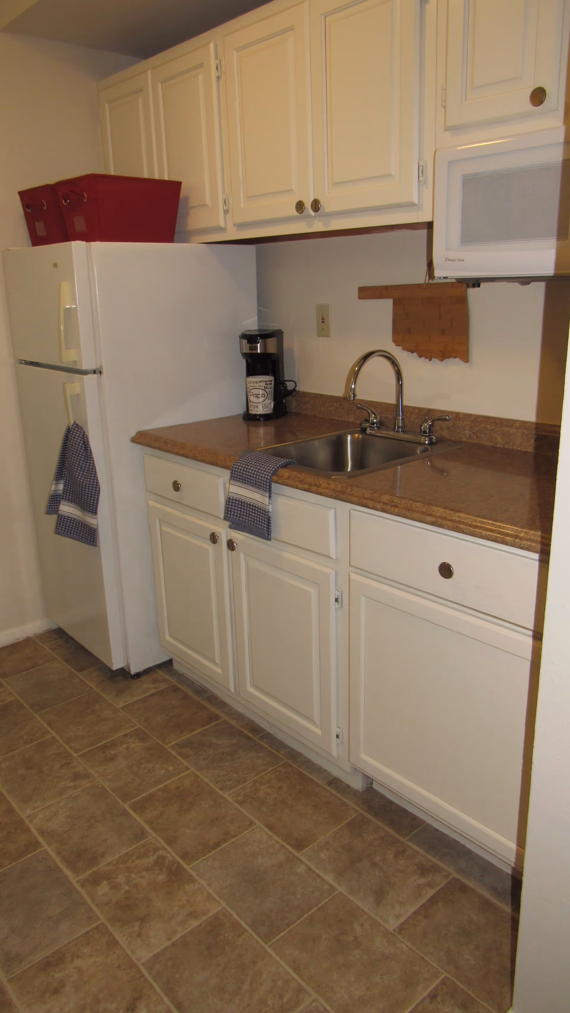 A small kitchen area with white cabinets, a brown countertop, a stainless steel sink with a faucet, a white refrigerator with a blue checkered towel hanging on the handle, a coffee maker on the counter, a microwave mounted above the counter, and two red storage bins on top of the refrigerator.