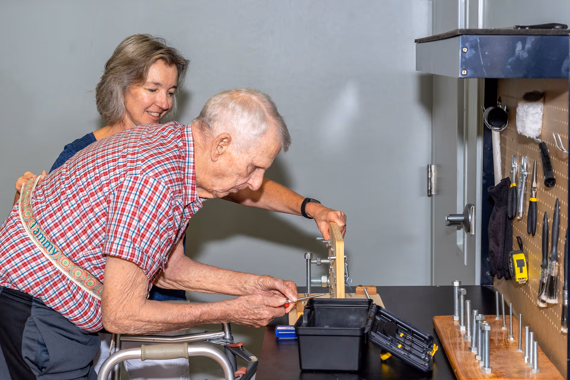 An elderly man using a screwdriver on a wooden piece held by a clamp on a workbench, assisted by a smiling woman standing behind him. The workbench has various tools and a pegboard with hanging tools in the background.