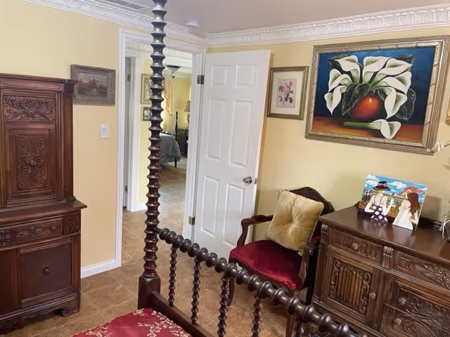 A cozy bedroom with antique wooden furniture including a bed with a carved headboard, a dresser, and a cabinet. The walls are painted yellow and decorated with framed paintings. A red cushioned chair with a beige pillow is placed next to the dresser. The door is open, showing a glimpse of another room with a bed.