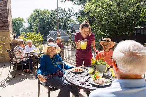 A group of elderly people sitting at outdoor tables enjoying food and drinks on a sunny day, with a caregiver serving lemonade. The setting is a patio area with trees and houses in the background.