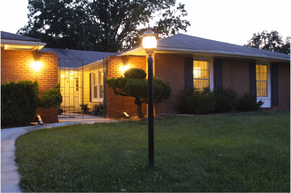 Exterior front of a single-story brick building at dusk with a lit lamppost, illuminated windows, walkway and shrubs.