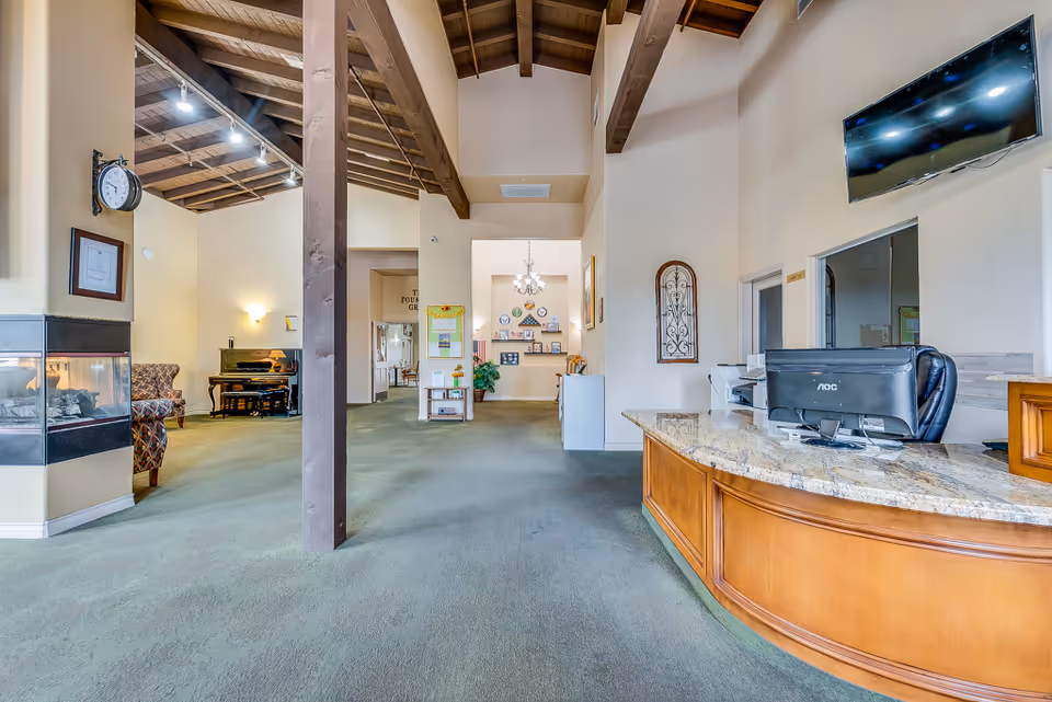 Spacious senior living reception area with a curved front desk, high exposed-beam ceiling, seating, piano, and fireplace.
