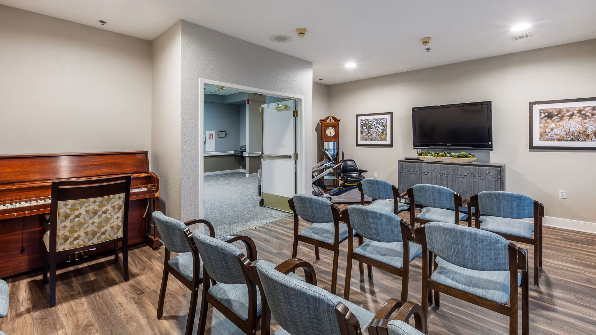 A cozy common room with several blue upholstered chairs arranged in rows facing a wall-mounted flat screen TV. To the left, there is a wooden piano with a chair in front of it. The room has wood flooring, neutral-colored walls, two framed pictures, and a grandfather clock near the TV. An open doorway leads to a carpeted hallway.