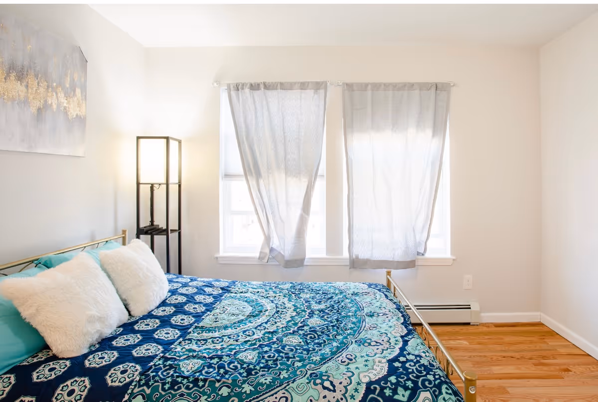 Sunlit bedroom with a patterned blue bedspread, two fluffy pillows, a floor lamp, and curtains over two windows.