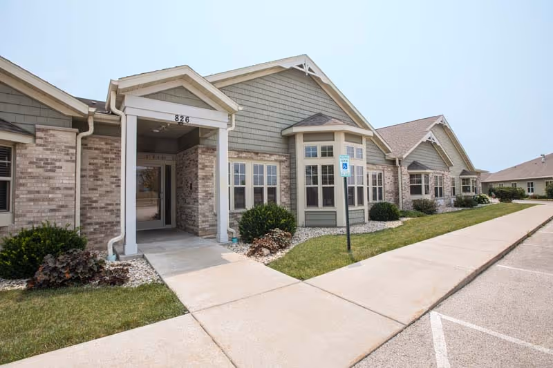 Exterior view of a single-story senior living facility building with brick and siding facade, multiple windows, a covered entrance with the number 826, landscaped bushes, and a sidewalk leading to the entrance. A handicap parking sign is visible near the entrance.