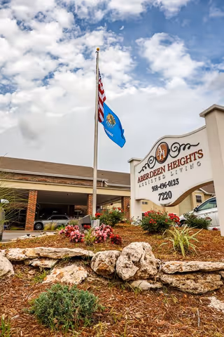 Exterior view of Aberdeen Heights Assisted Living facility with a sign displaying the facility name, phone number, and address. There is a flagpole with the American flag and another flag, surrounded by landscaped rocks and flowers under a partly cloudy sky.