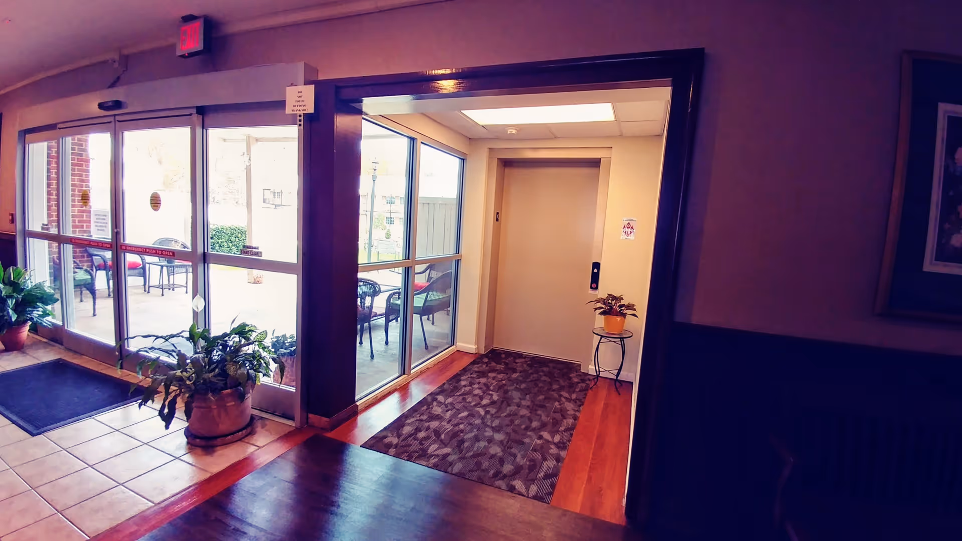 Interior view of an entrance area in a senior living facility showing glass sliding doors leading outside to a patio with chairs and tables. There is a potted plant near the door and an elevator with a small table and plant beside it inside the building. The floor has a combination of tile, wood, and carpet.