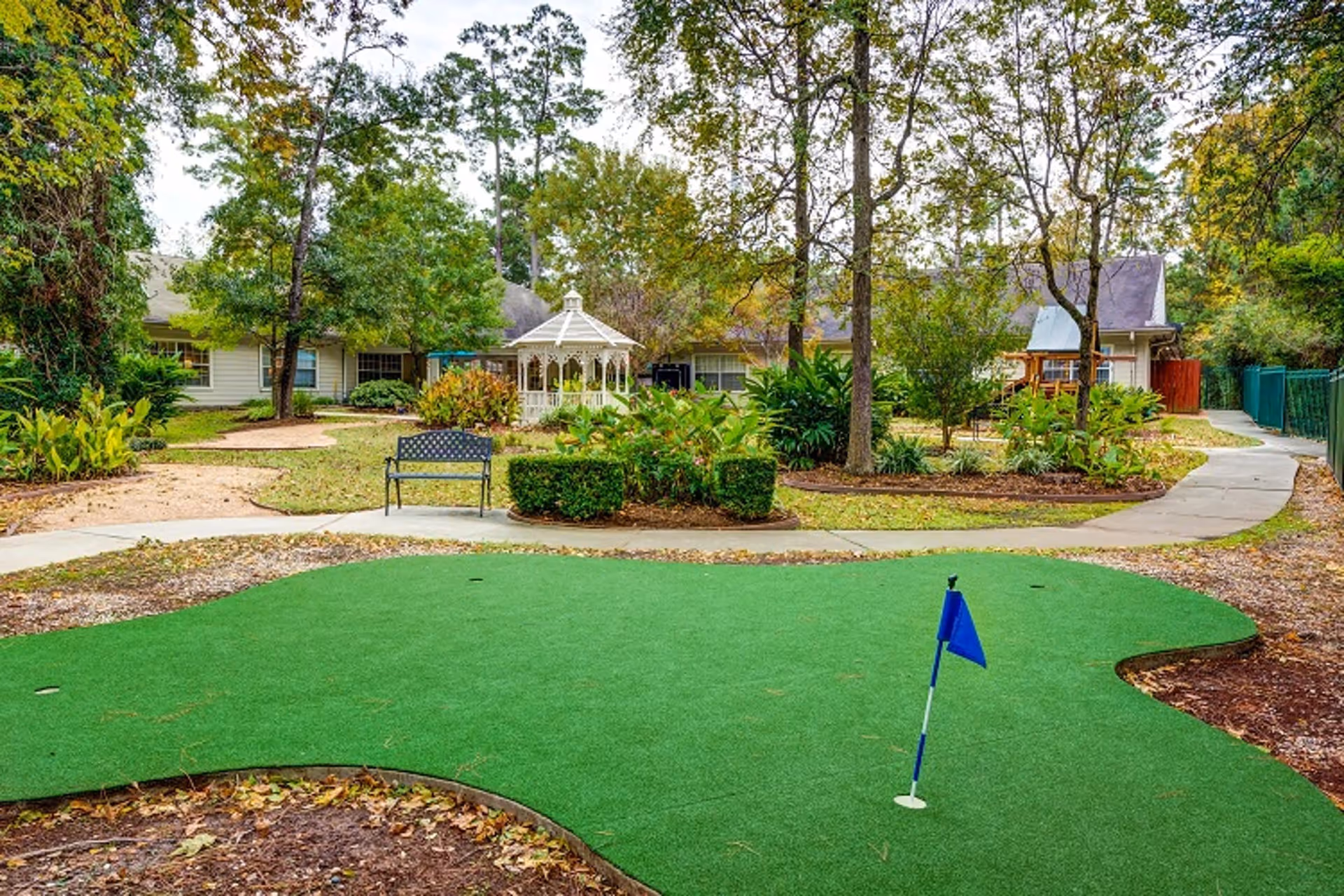 Outdoor garden area with a putting green featuring a blue flag, surrounded by trees, bushes, and a paved walkway. In the background, there is a white gazebo and residential-style buildings.