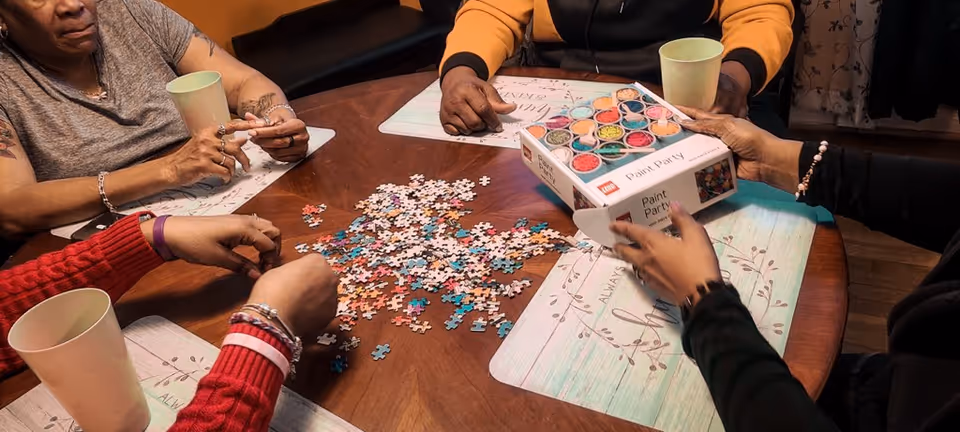 Four people sitting around a wooden table working on a colorful jigsaw puzzle. One person is holding a box labeled 'Paint Party' with images of paint containers on it. There are placemats and cups on the table.