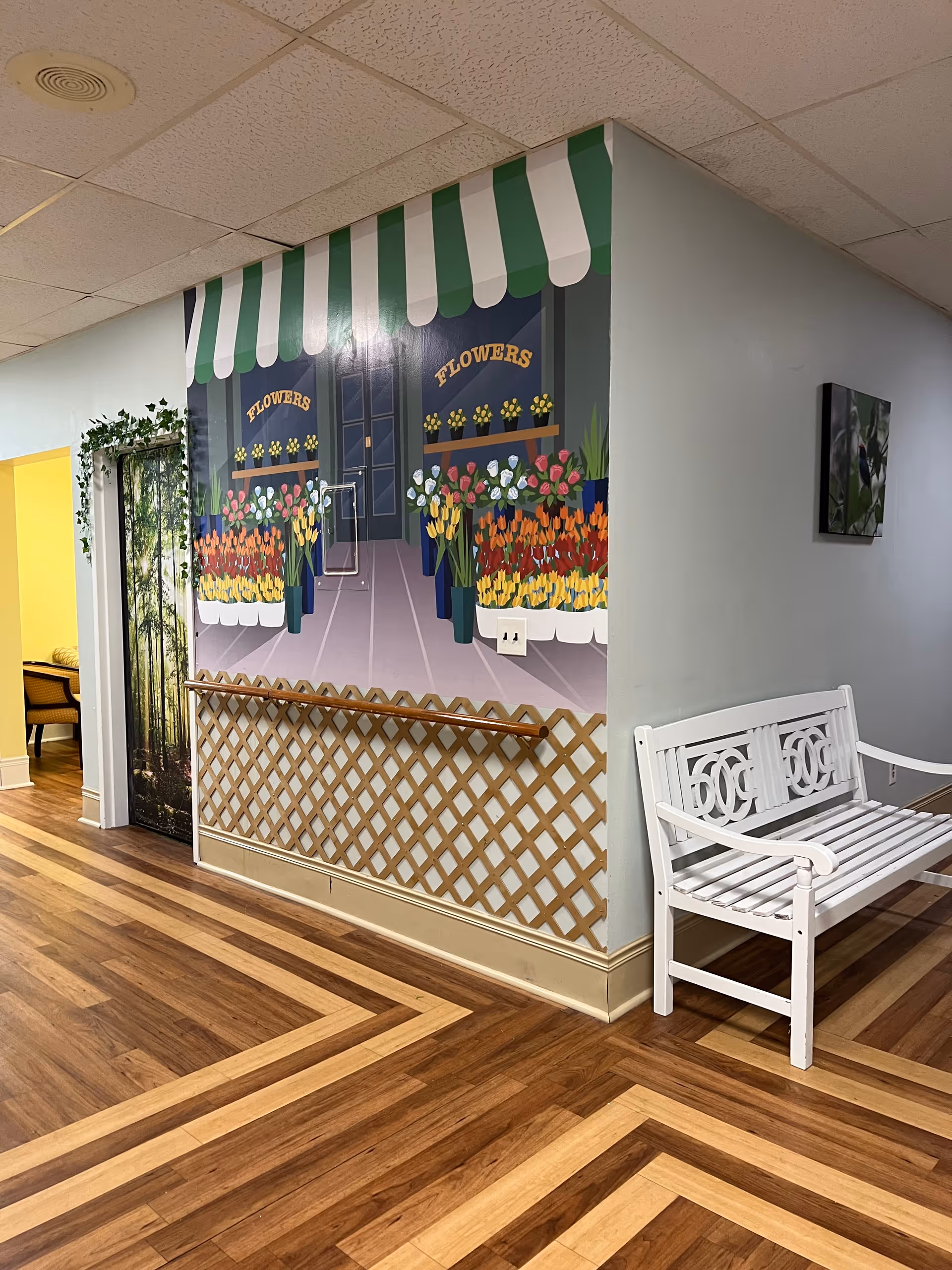 Interior hallway of a senior living community with a mural of a flower shop on the wall, featuring colorful flowers under a green and white striped awning. There is a white wooden bench on the right side and a handrail along the wall beneath the mural. The floor has a patterned wood design, and there is a doorway with a nature-themed curtain on the left.