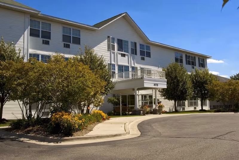 Front exterior of a three‑story white senior living building with a covered entrance, landscaping, and a circular driveway under a blue sky.