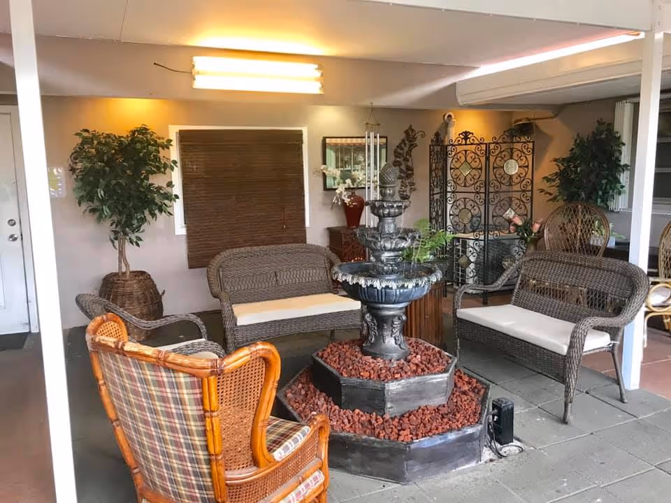 Indoor seating area with wicker chairs and a loveseat arranged around a decorative black fountain filled with red rocks. The room has potted plants, a decorative screen, and a window with a brown blind. The lighting is warm and the floor is tiled.