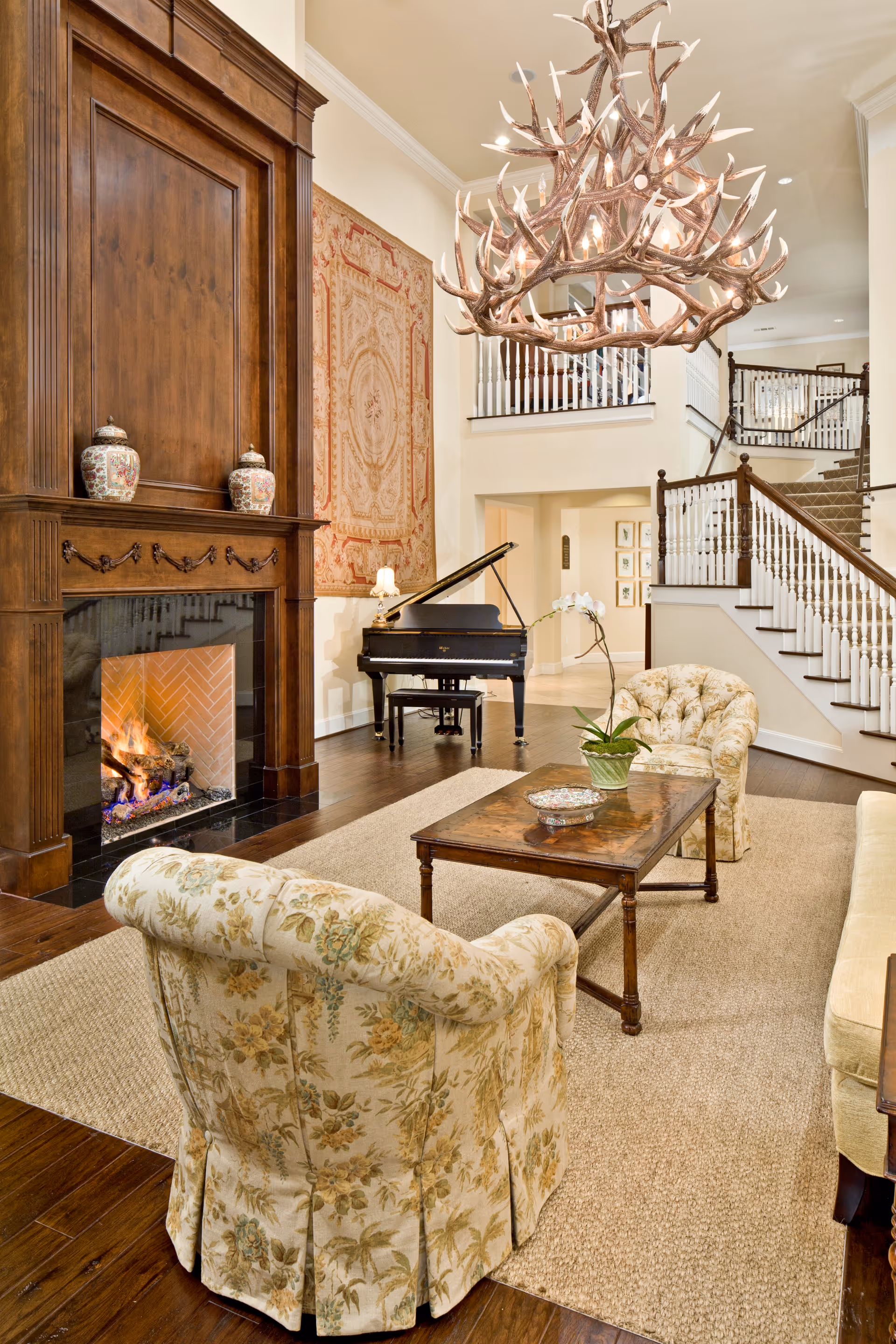 A cozy living room with a large wooden fireplace on the left, a lit fire inside. Two floral upholstered armchairs and a wooden coffee table with a plant and decorative bowl are placed on a beige rug. A grand piano is positioned near the back wall with a decorative tapestry hanging above it. A large antler chandelier hangs from the ceiling, and a staircase with white railings and dark wood banisters is visible in the background.